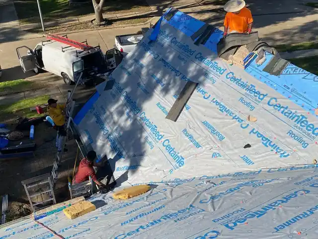 Integrity Roofing Pros crew rolling CertainTeed RoofRunner underlayment across a roof deck, work van and ladders at the curb below