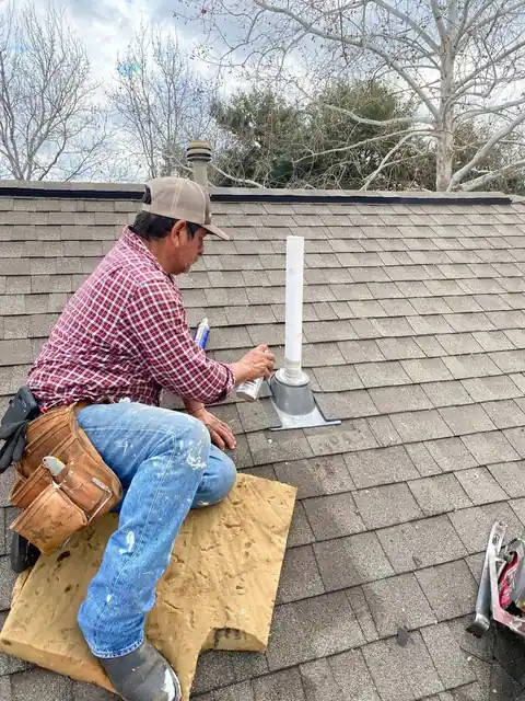 Roofer kneeling on a shingle roof sealing a plumbing vent boot
