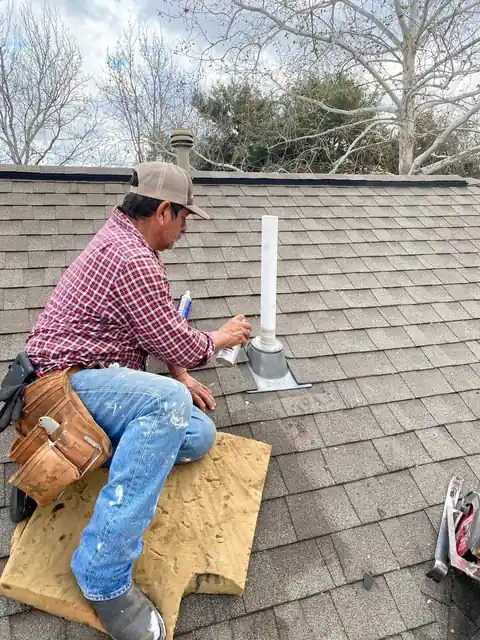 Roofer in a plaid shirt sealing a plumbing vent flashing boot on shingles
